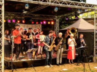 Foto: Orchester mit vielen Blasinstrumenten spielt teilweise auf einer B&uuml;hne und davor
