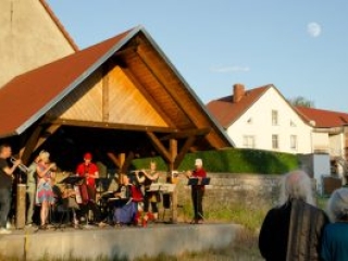 Foto: Musikgruppe spielt unter einem offenen Holzdach. Ehepaar im Vordergrund schaut zu und der Mond steht am Himmel.