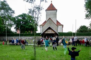 Foto: Junges und altes Publikum tanzend auf einer grünen Wiese. Im Hintergrund sitzen viele Menschen und lauschen dem Orchester auf der überdachten Bühne, vor einer Kirche.