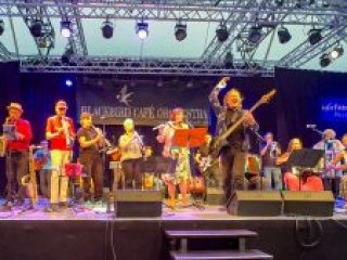 Foto: Orchester mit verschiedenen Instrumenten auf einer B&uuml;hne, im Vordergrund Mann mit Bassgitarre und einer erhobenen Hand