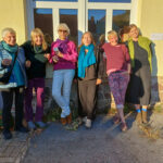 Foto: 6 Frauen lehnen mit dem Rücken an einer Wand mit Fenster und lächeln